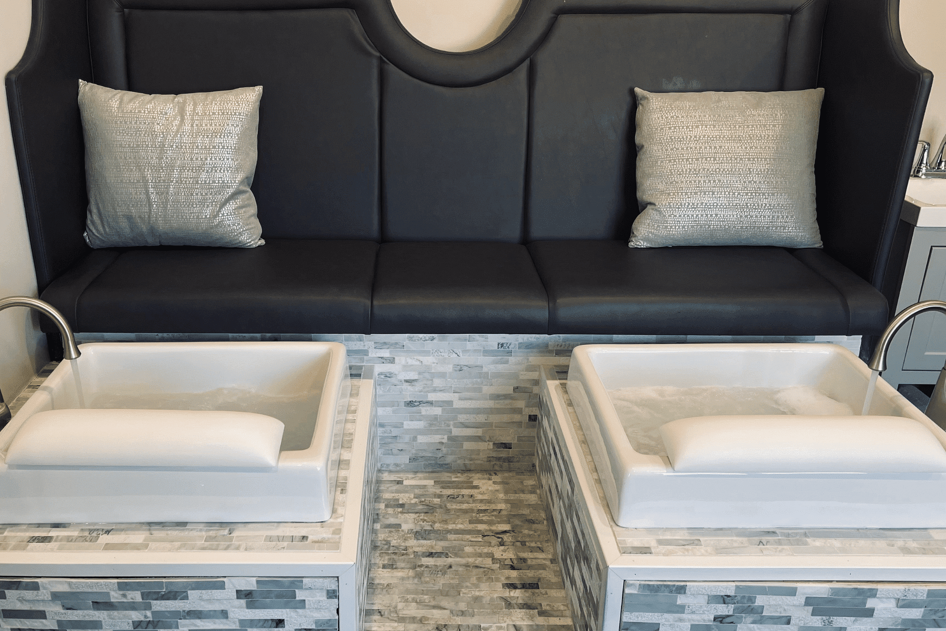 Modern pedicure station with cushioned bench and two foot spas in a sleek, tiled setting.